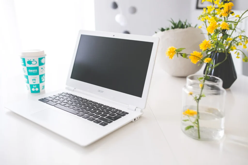 Weißer Laptop auf einem hellen Schreibtisch mit Kaffee und gelben Blumen