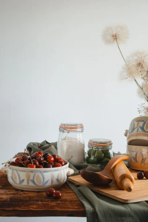 Schale mit Kirschen, Vorratsgläser und Backutensilien auf einem Holztisch